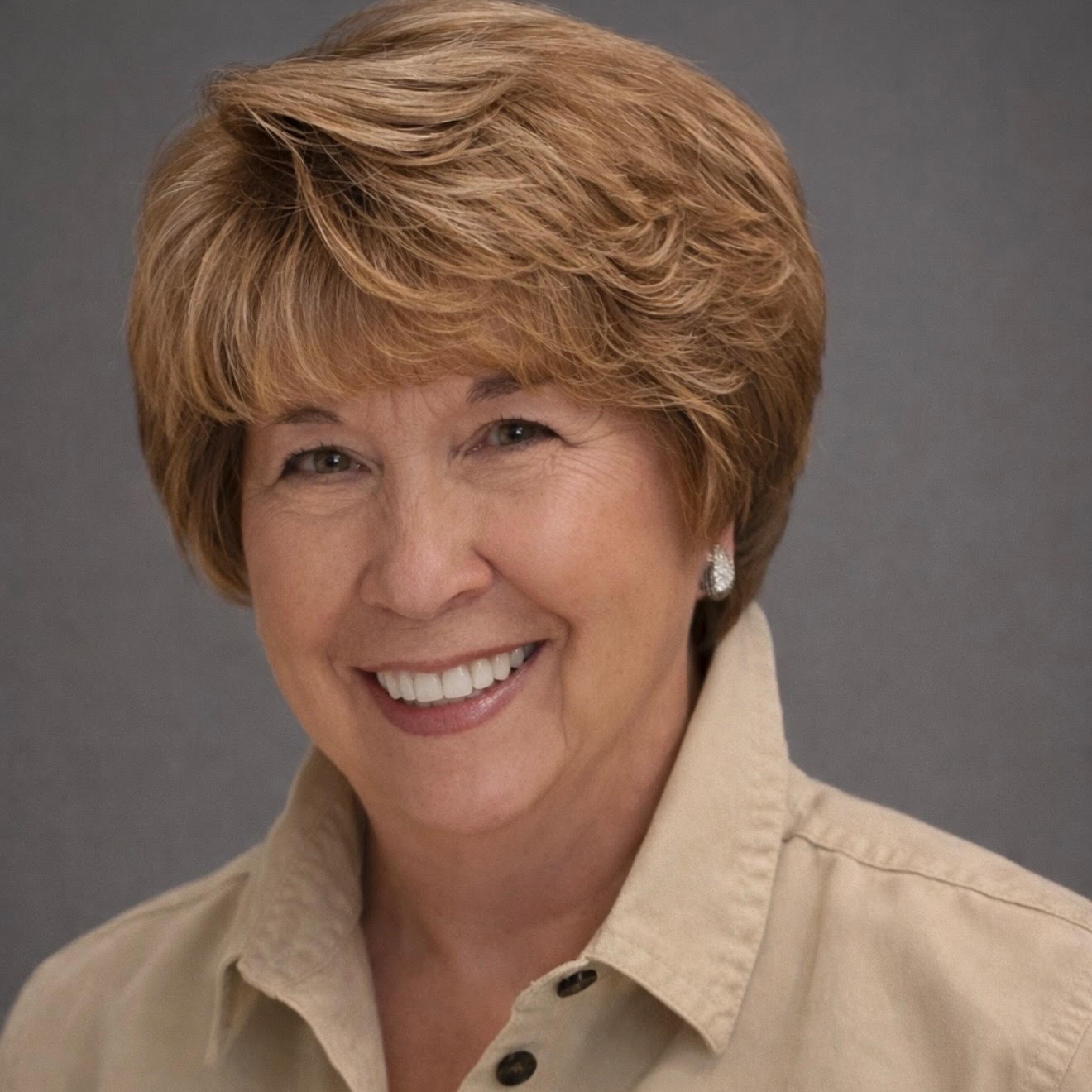 Smiling woman with short, light brown hair wearing a beige collared shirt and earrings, set against a gray background.
